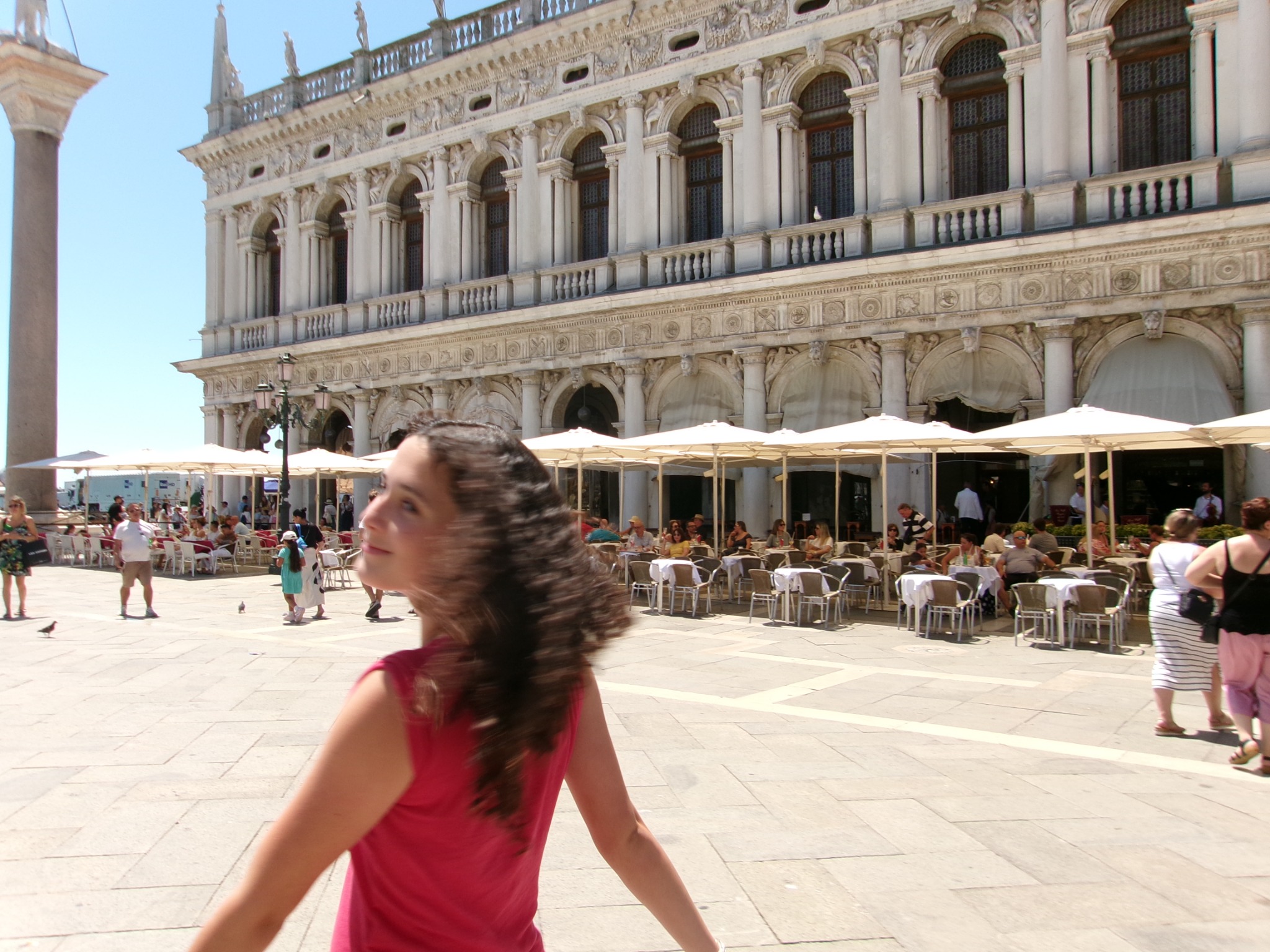 A person in red turning in a bright square beside an ornate building